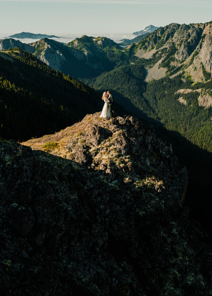 A  shot of a couple in their wedding attire in a mountain landscape as they celebrate their Olympic National Park elopement. The sun washes over the landscape with golden light and shadows slice through the landscape 