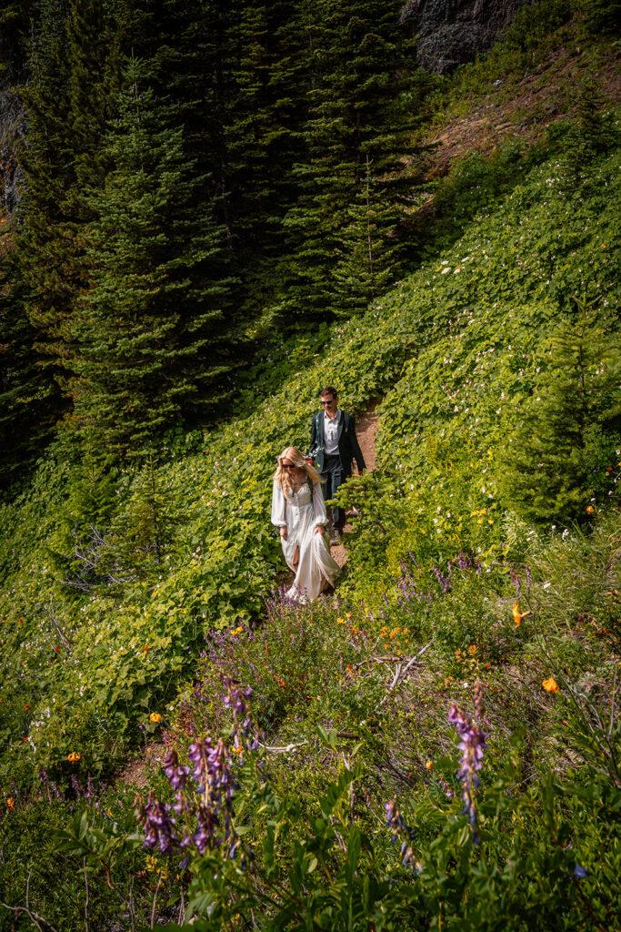 a bride and groom descend down a wildflower framed switchback in their wedding attire and hiking bags during their Olympic National Park elopement