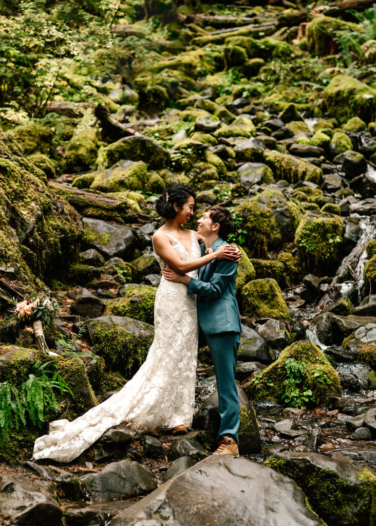 a couple embraces a top mossy rocks, smiling at each other during their Olympic National Park wedding 