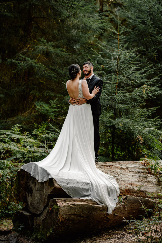 A couple in their wedding attire embrace on top of a massive log in the middle of a green forest. The brides dress cascades down as her groom smiles widely at her 