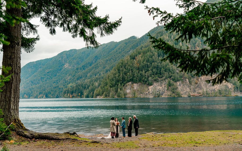 A couple and their guests share their Olympic National Park elopement with a small group of guests on the shores of lake crescent. the blue of the lake feels brilliant and sparkling.
