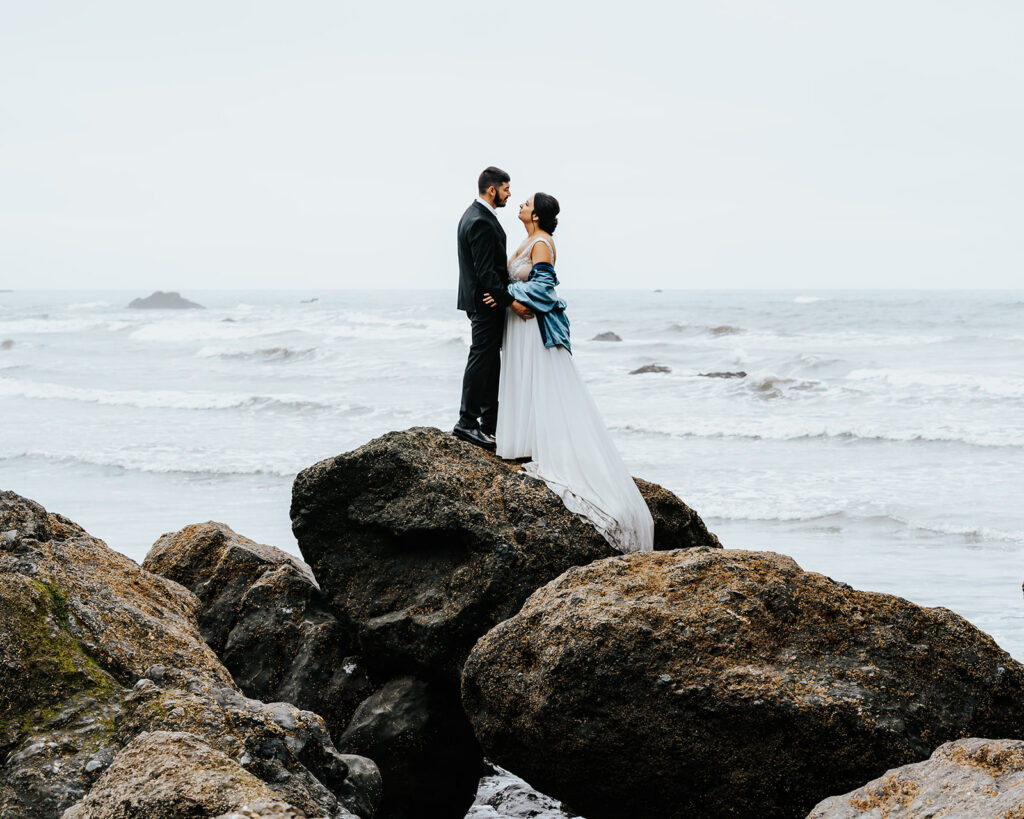 A couple in their wedding attire embrace on a series of boulders. waves crash behind them as they embrace on the rocky beach during their Olympic National Park elopement