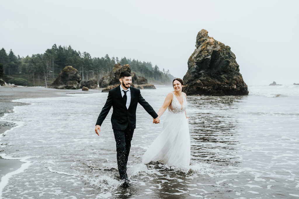 a bride smiling and laughing widely at her groom as they run through the waves on a rocky beach 
