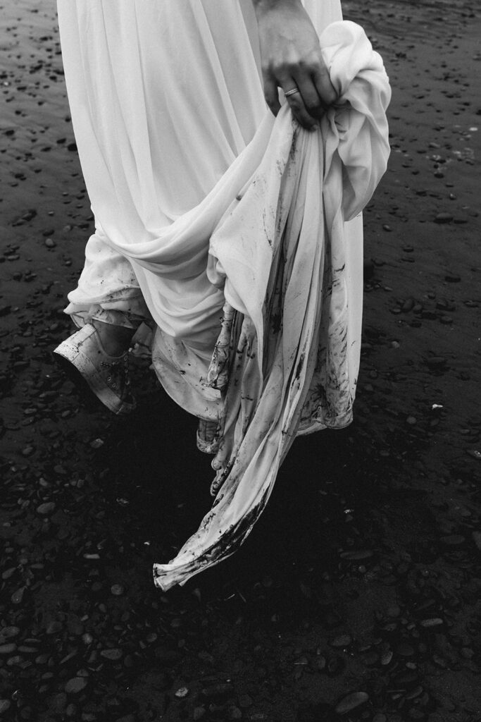 a black and white image of a sopping wet wedding dress. the ends of it dragging through the muddy shore of the beach 