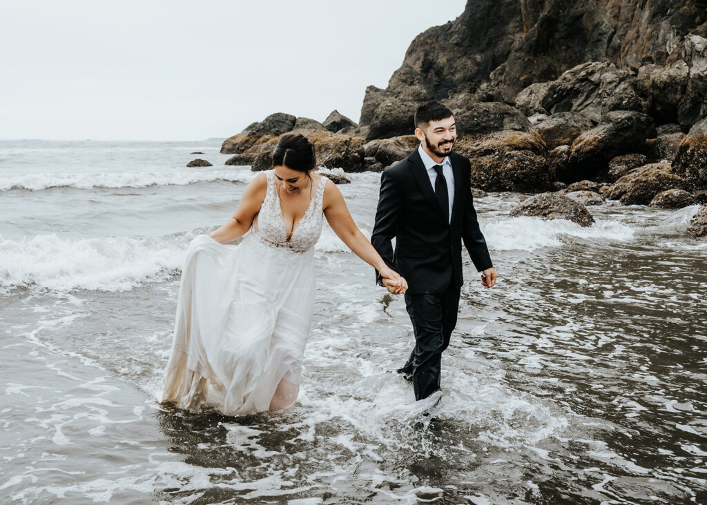 A couple in their wedding attire splash through the icy waves of the Pacific Ocean. They smile widely as they romp around during their Olympic National Park elopement