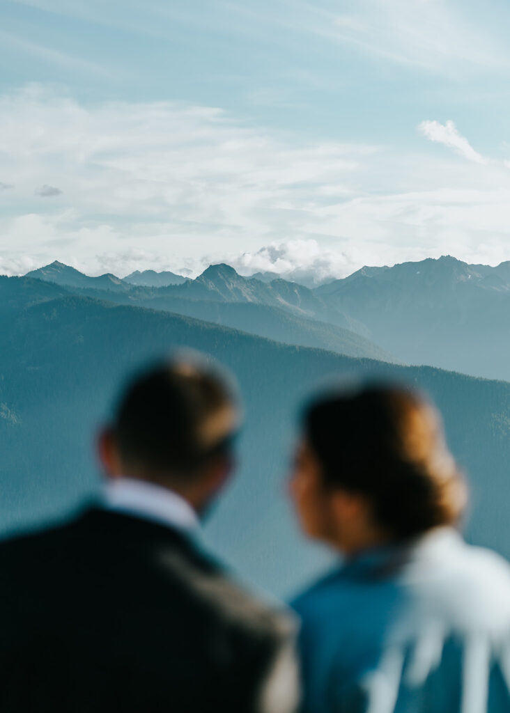 The soft profile of a couple looking out at the Olympic Mountains. The mountains are blue during sunset and fluffy clouds billow behind them during their Olympic National Park elopement