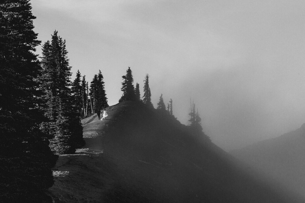 a bride and groom explore up a mountains edge. the landscape is contrasted by sunshine on one side, and thick fog on the other. 
