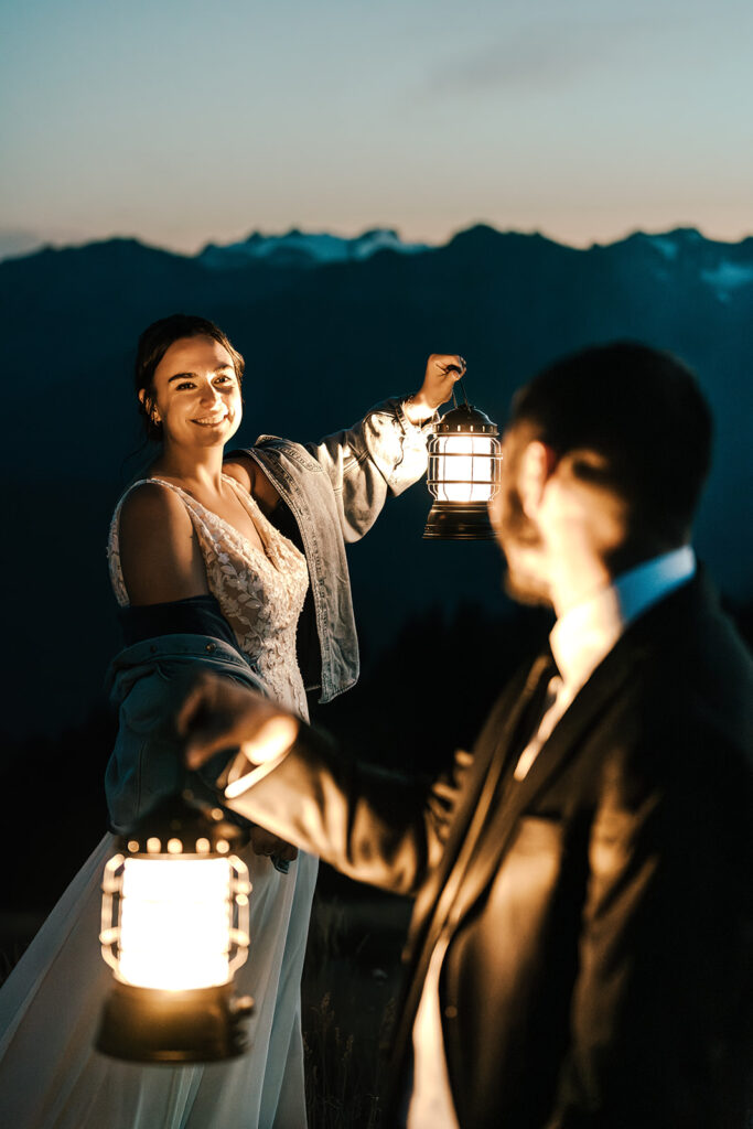 a bride is lit by lantern light as her groom gazes lovingly at her. She is framed by him and his lantern. Behind her, we see snowy mountains in the dusky blue hour of their Olympic National Park elopement