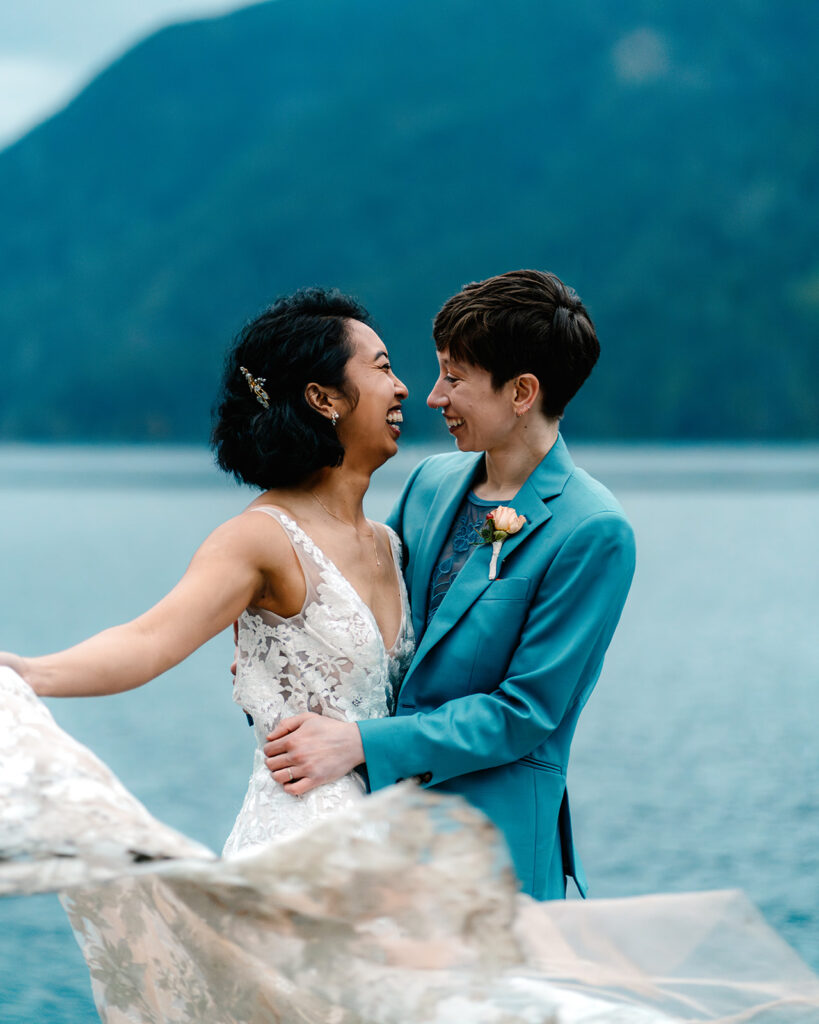 two brides smile widely at each other as they dance by a bright blue lake 