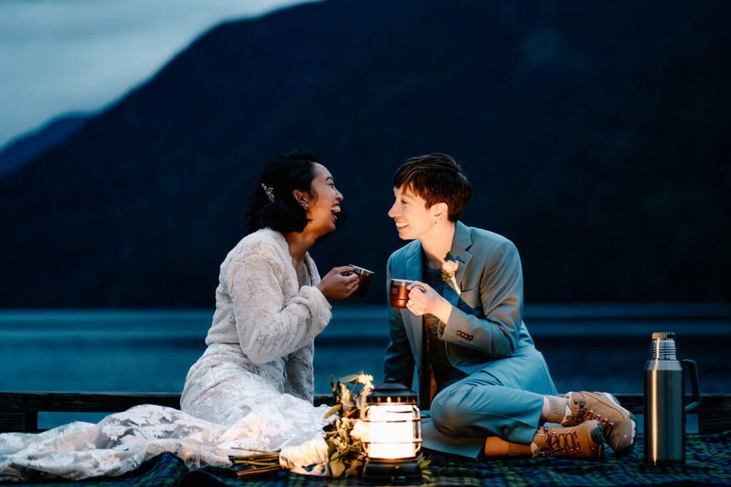 Two brides smile and laugh as they share tea at the edge of a dock on lake crescent during blue hour to end their Olympic National Park elopement.