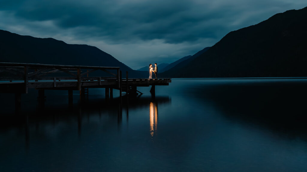 a shot taken at blue hour. a couple stands on the edge of a dock. lit only by lntern light, we see the glassy reflection in the still waters below them 