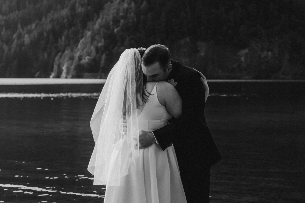 a black and white image of a bride and groom embracing during their Olympic National Park wedding. the moment is tender, and ew see rays of light sparkling on the lake behind them 