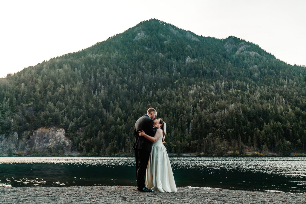 A couple in their wedding attire embrace on the shore of a sparkling lake. Green mountains peak behind them as they kiss during their Olympic National Park elopement