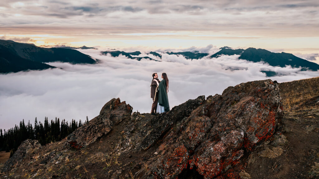 A couple in their wedding attire and wearing cloaks that suggest middle earth, or a more ethereal vibe embrace above a stunning cloud inversion during their Olympic National Park elopement