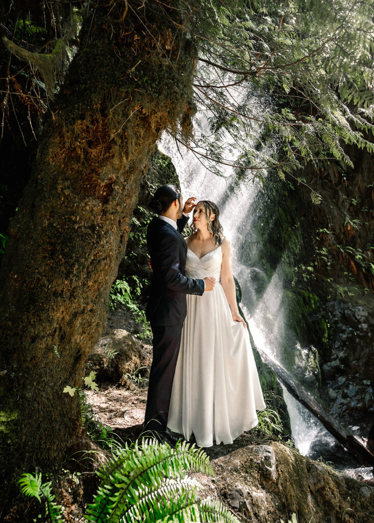 A couple in wedding attire gaze lovingly at each other in front of a sparkling waterfall. light shines down on the water creating a dazzling effect during their elopement day
