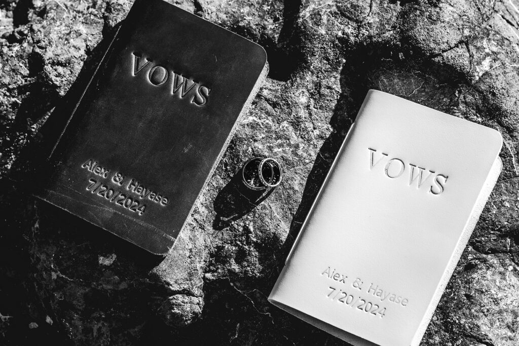 a black and white photo of wedding bands and vow books resting on a rock as sunlight creates harsh shadows 