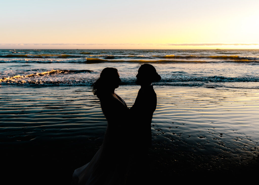 the dark silhouettes of a couple are highlighted against the colorful rolling waves of the Pacific highlighted by a golden sunset during their olympic national park wedding 