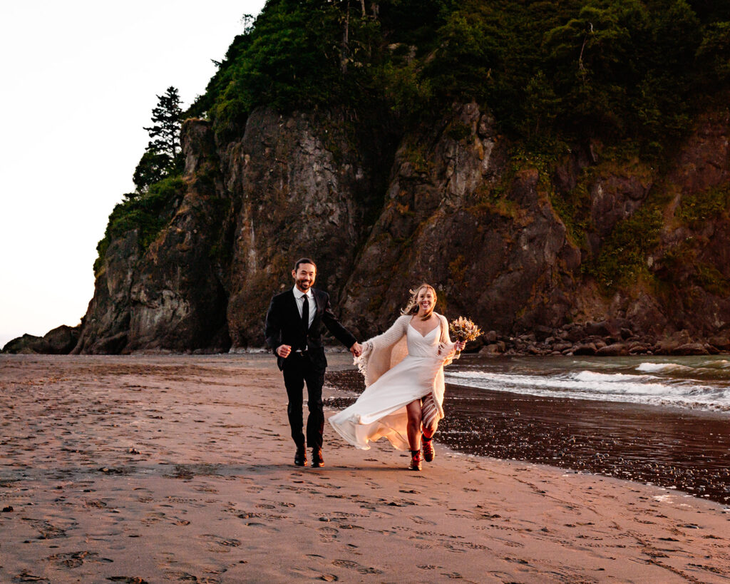a bride and groom run down a rocky beach. Warm, golden light from the sunset highlights them as they play