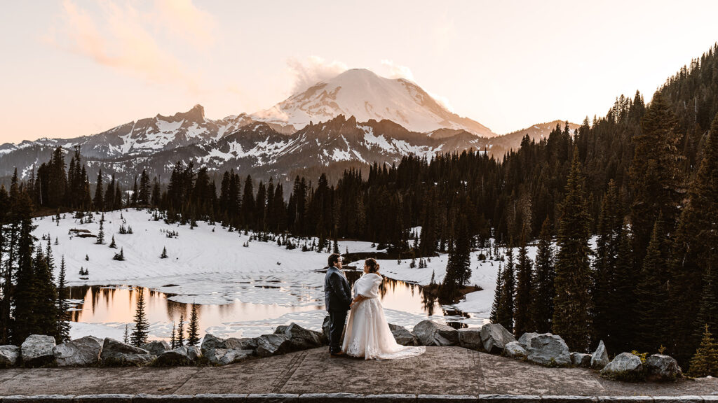 a couple in their wedding attire embrace in a snowy landscape. Mount Rainier in highlighted by the sunset behind them in one of the best places to elope in Washington