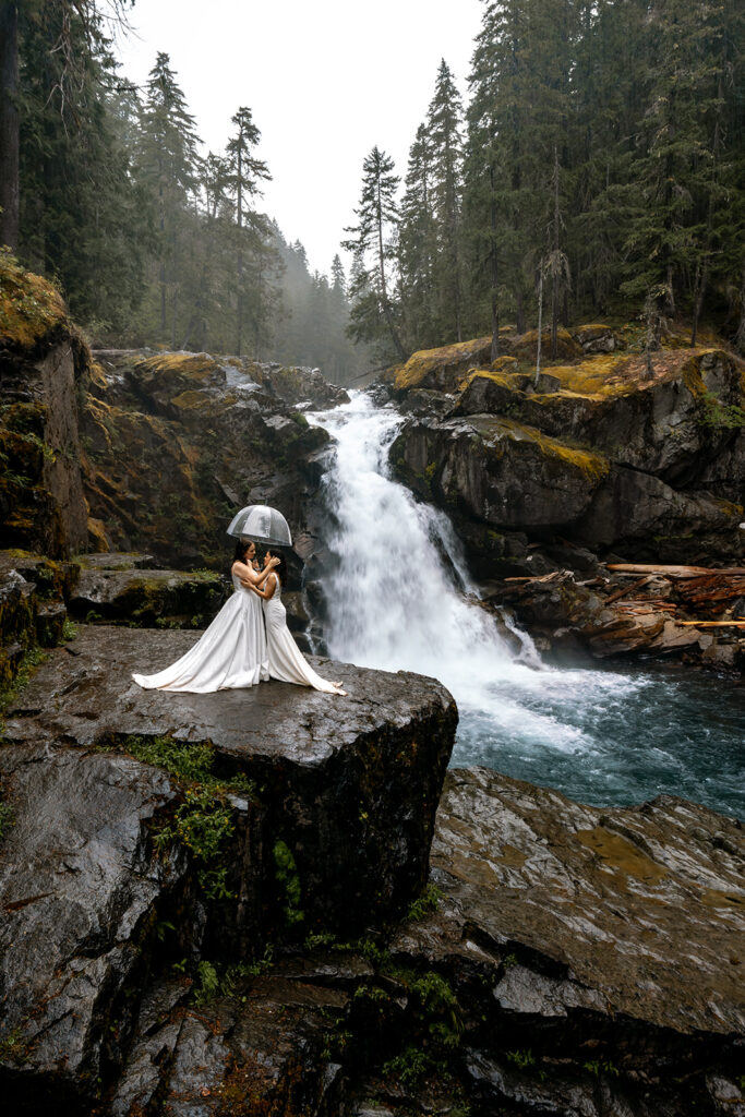 Two brides embrace under a clear umbrella as they explore a striking turquoise waterfall in mount rainier one of the best places to elope in Washington