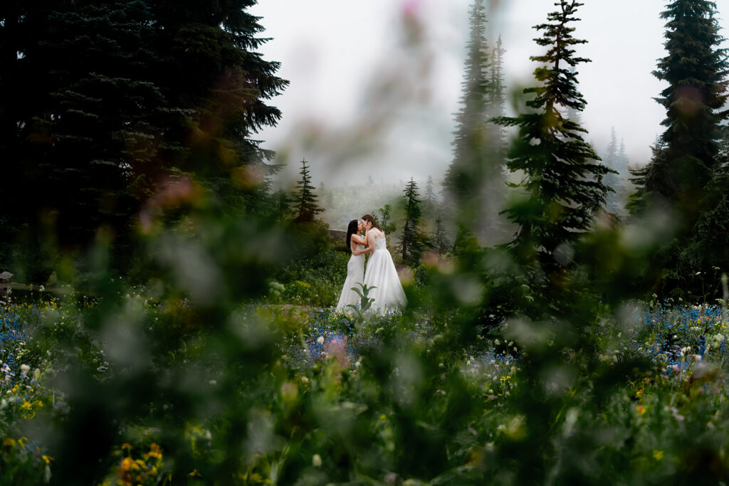 framed by wildflowers, two brides kiss as they immerse themselves in one of the best places to elope in Washington. Colorful flowers and swirling, white fog create the feeling that you're looking at an oil painting instead of a photograph 