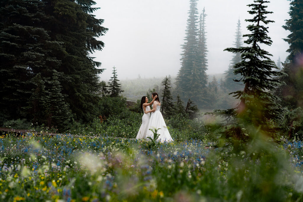 two brides embrace in a meadow dotted with sprays of wildflowers. The atmosphere is ethereal, as fog swirls around them while they explore one of the best places to elope in Washington