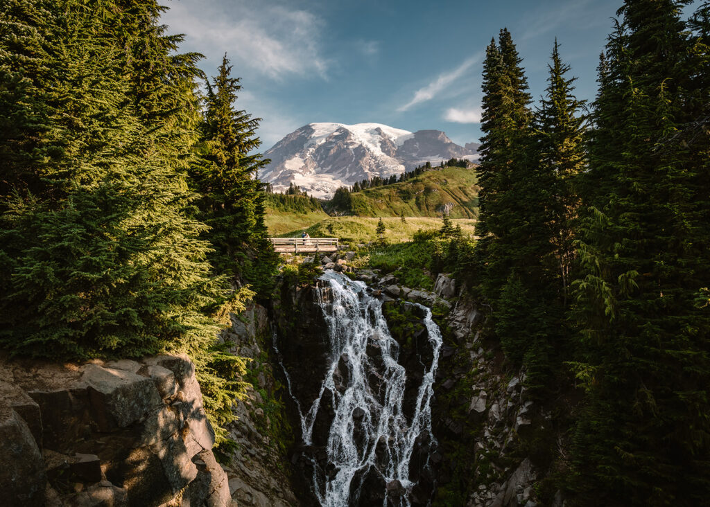 a couple in their wedding attire embrace on a bridge over a waterfall. Mount Rainier towers behind them as they embrace in one of the best places to elope in Washington