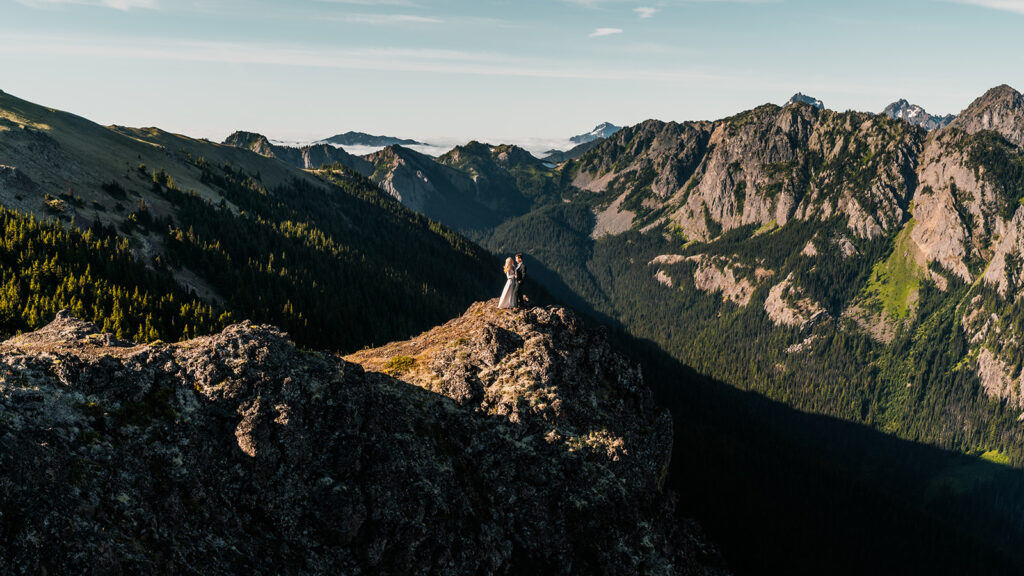 a bride and groom stand on the peak of a rocky outcropping in their wedding attire. Sunlight slices across the landscape and highlights them as they immerse themselves in one of the best places to elope in Washington
