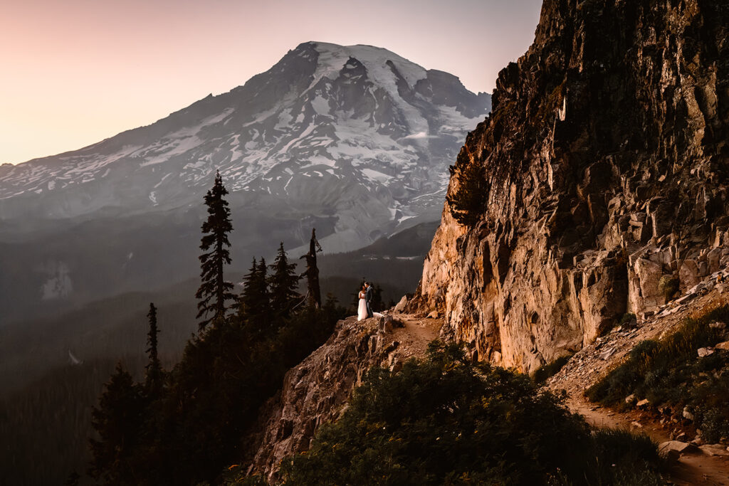 a wide landscape shot at dusk with Mount Rainier towering in the background. A couples, perfectly centered with the peak of the mountain and the winding path drawing our eye, embrace in their wedding attire in one of the best places to elope in Washington