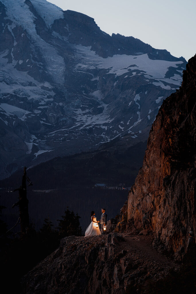 a couple stand on a trail in their wedding attire during blue hour. They are lit only by the final wash of slight sunlight and lanterns.