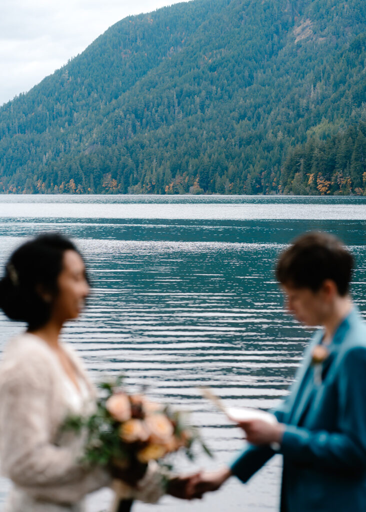 a couple in their wedding attire create the frame of this image, focused on the turquoise waters of Lake Crescent behind them as they exchange vows
