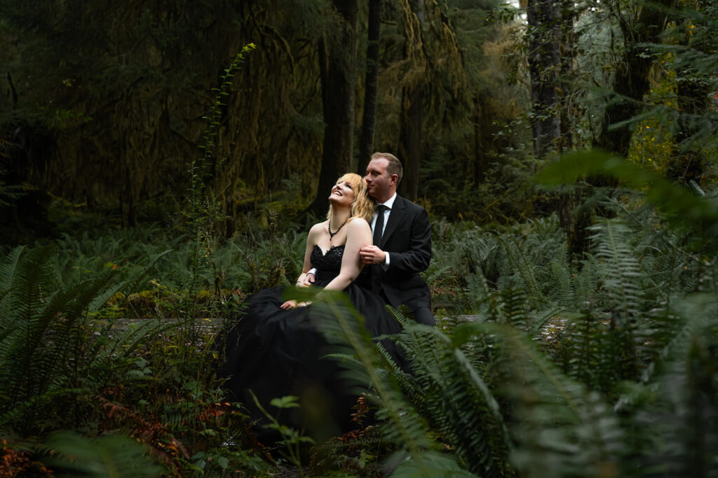 A couple in all black wedding attire embrace in a lush, green forest. They are surrounded by verdant ferns and hanging moss as they immerse themselves in one of the best places to elope in Washington