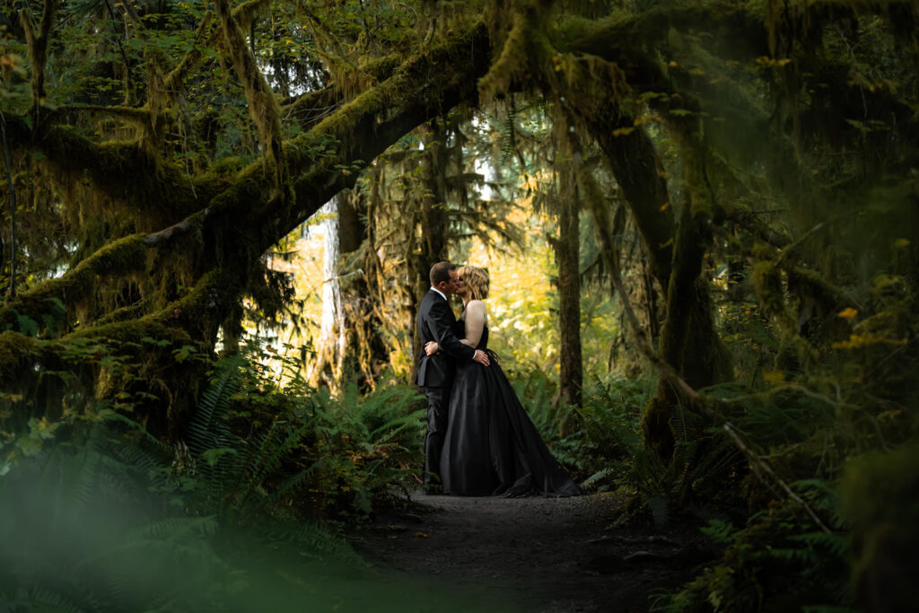 a couple in all black wedding attire embrace under the lush, green canopy of the Hoh Rainforest.