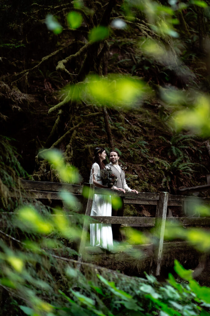 a couple in their wedding attire gaze lovingly at each other while standing on a wooden bridge. They are framed by the lush greenery of the Olympics, one of the best places to elope in Washington