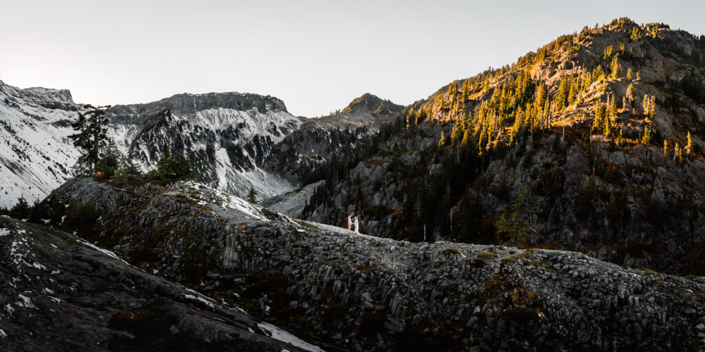 A large landscape showing off mountains dusted with early snow, and golden sunlight hitting the eastern peaks. In the center, we see a tiny couple in wedding attire exchanging vows in one of the best places to elope in Washington.
