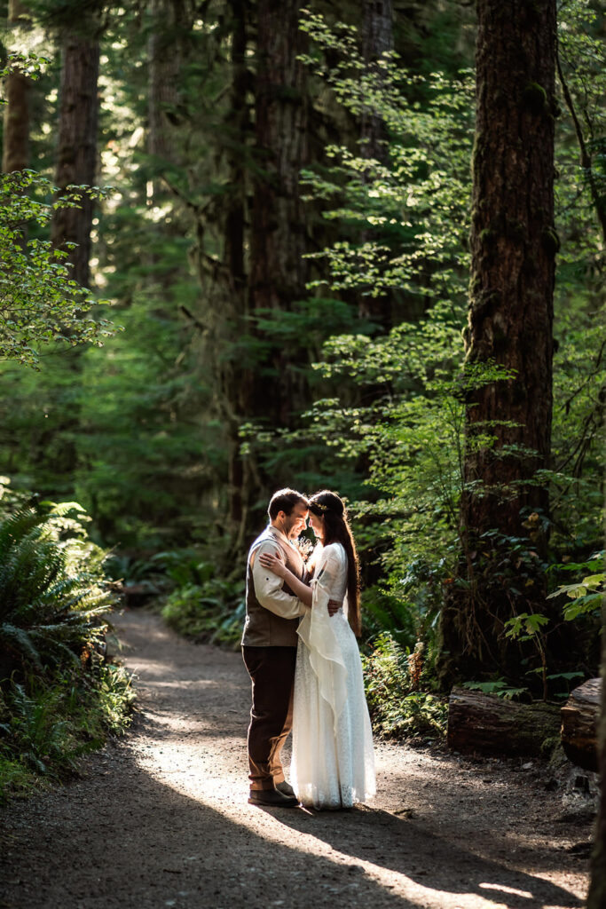 A couple in wedding attire embrace in golden light filtered through a dense, green canopy. The forest surrounds them on their wedding day as they explore one of the best places to elope in Washington