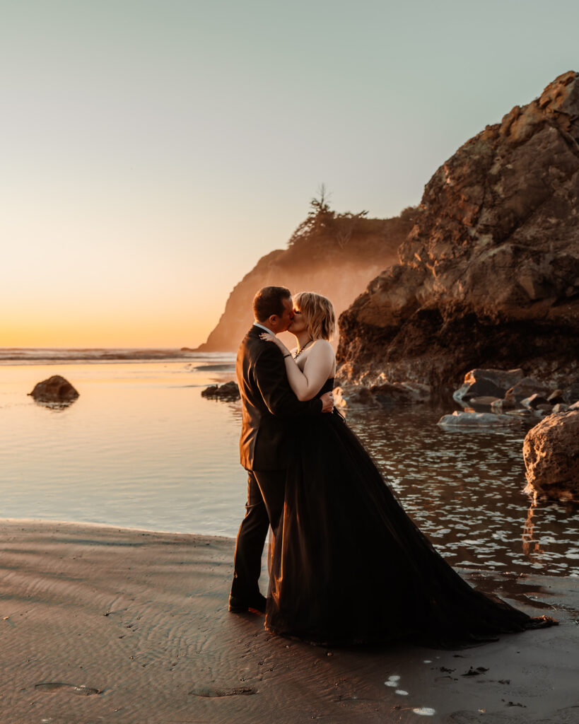 A couple in all black wedding attire kiss on a rocky beach. The punchy colors of the sunset glow around them and highlight the moment as they explore one of the best places to elope in Washington