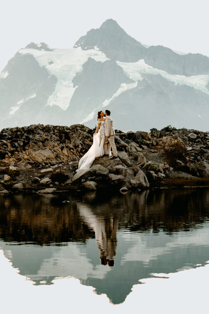 pale, blue light surrounds a couple in their wedding attire as they kiss by a reflective tarn. The morning light is soft as they explore one of the best places to elope in Washington