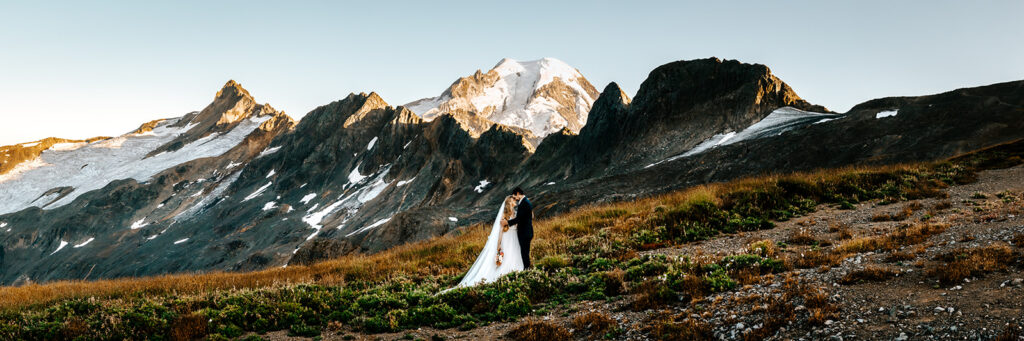 a panoramic shot of a couple in wedding attire embracing in front of mount Baker. The mountain is so close, you feel like you could touch it  making it truly one of the best places to elope in Washington