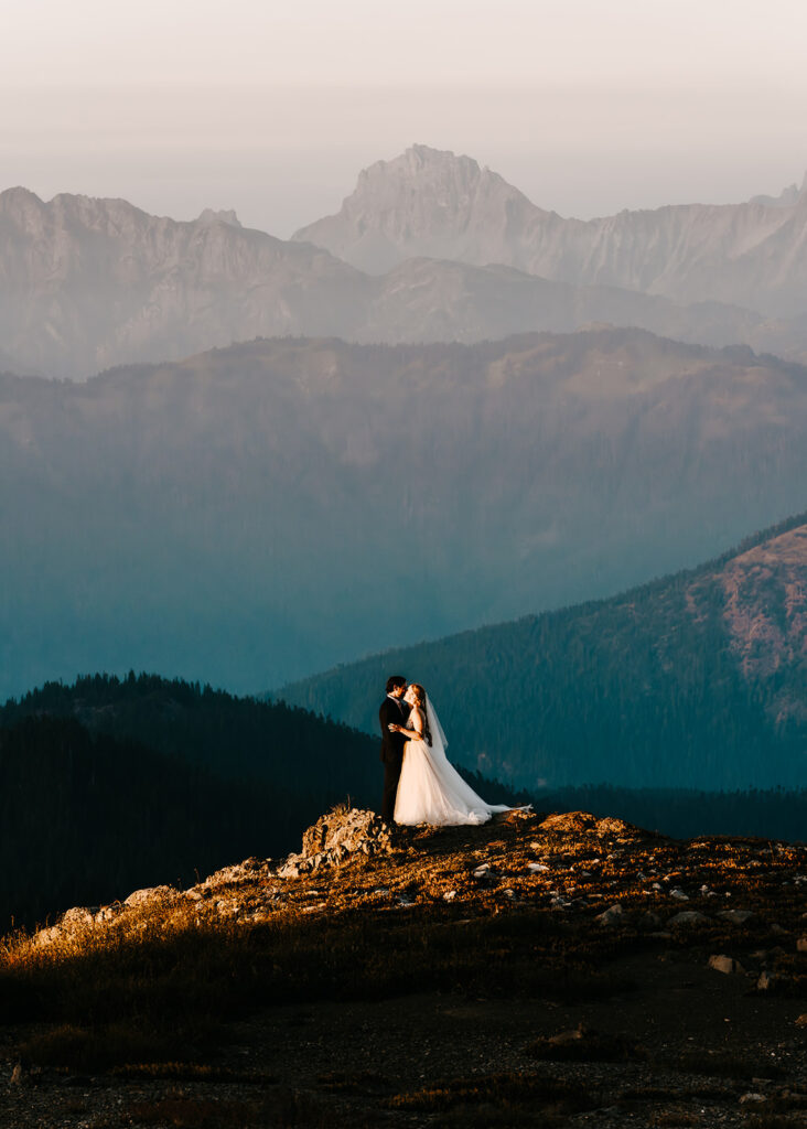 A couple, in their wedding attire embrace on the edge of a mountian. The layers of the skagit range roll behind them and fade from a deep teal to a dusty purple in one of the best places to elope in Washington