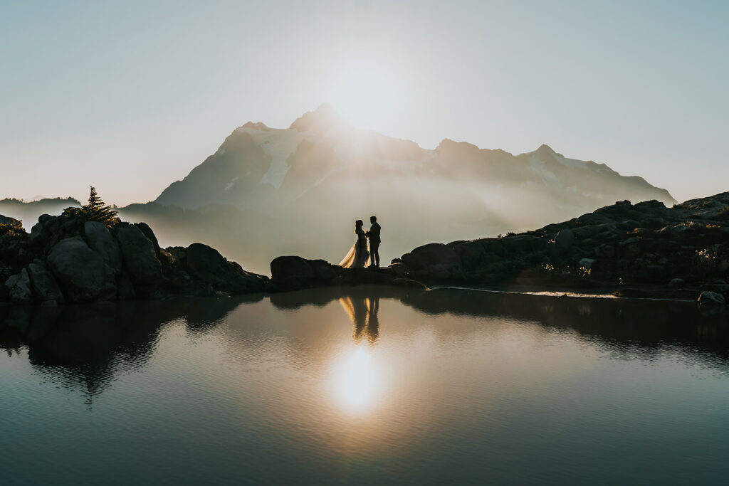 a couple in wedding attire stand centered on top of a reflective tarn. Mountains rise behind them as does the sun in one of the best places to elope in Washington