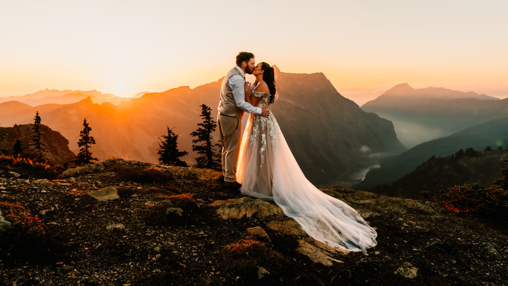 a couple embrace on the top of a mountain in their wedding attire. Mountains and lakes roll behind and beneath them as they explore one of the best places to elope in Washington
