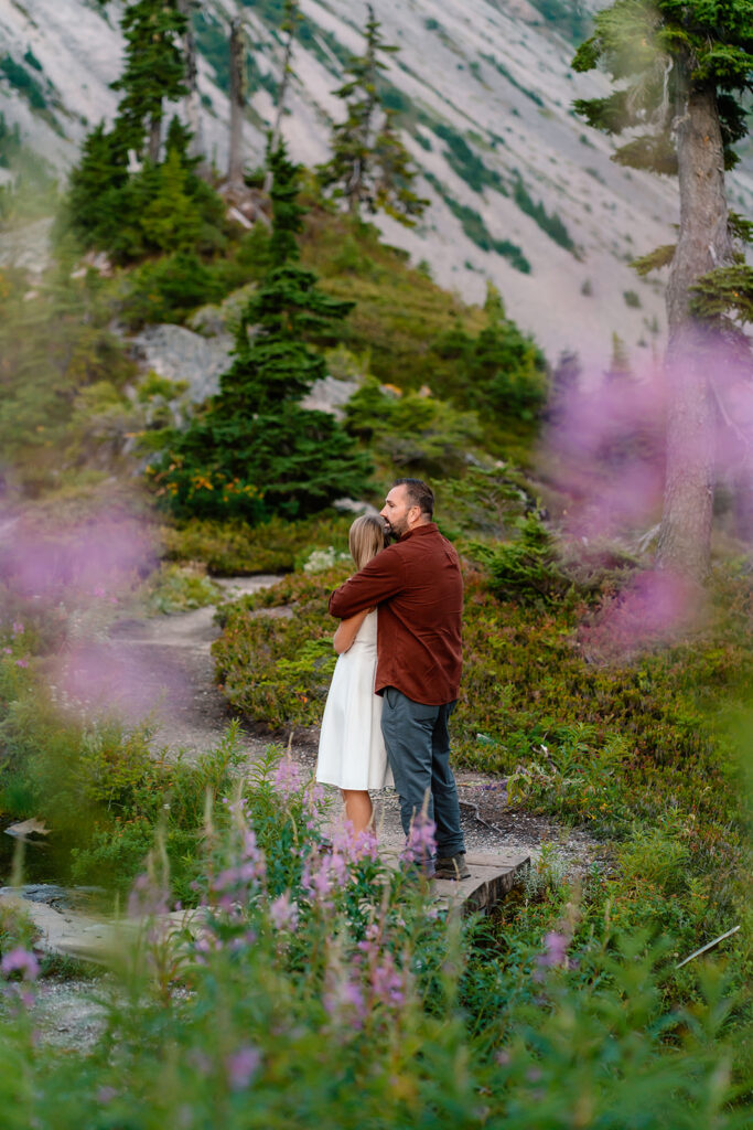 a bride gazes out at the landscape as her groom wraps his arms around her. they are framed by blurry, purple Alaskan fireweed.