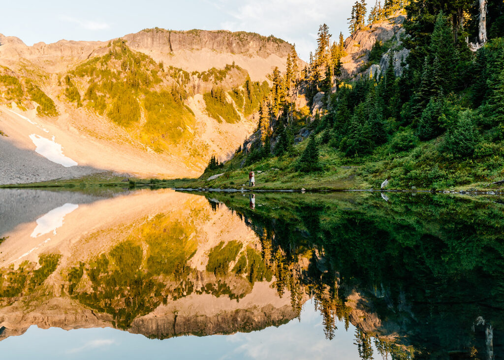 a tiny couple in a massive landscape. We see them embrace in a meadow on the edge of a stunning reflective lake. behind them we see table mountain with a stunning alpine glow. we see this reflected in the still waters below them on the morning of their mt baker micro wedding 