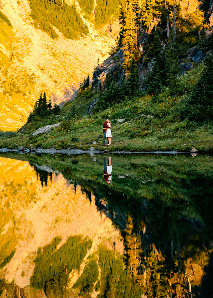 a vertical shot of  a stunning sunrise scene by a reflective lake. we see a bride and groom in casual wedding attire embrace. Green wildflower meadows rise above them, and a glowing mountainside serves as the background. The scenery is then reflected back to us in the still waters of the lake below them  