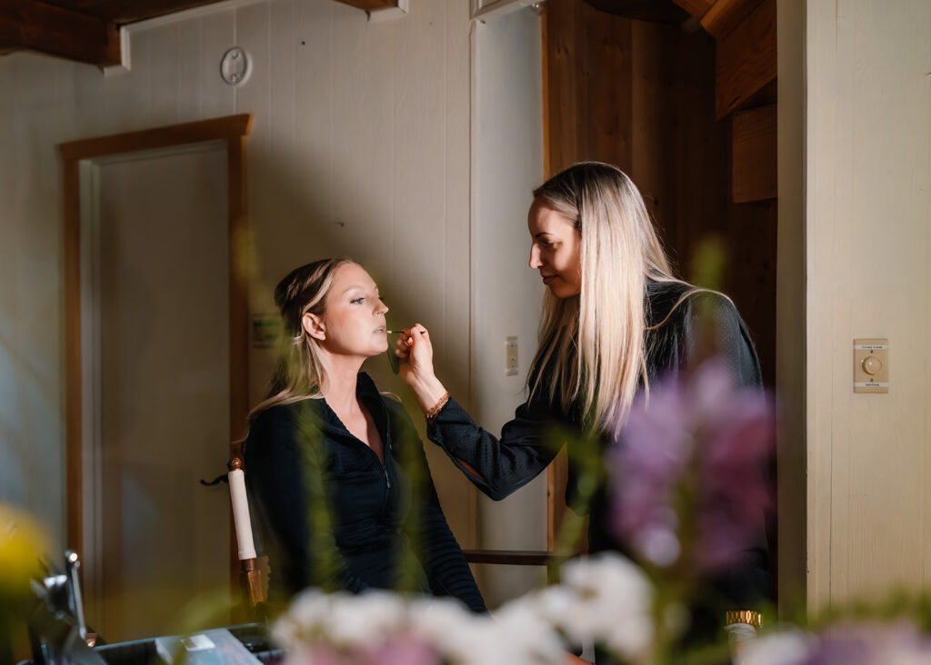 shot through a bridal bouquet, we see a bride getting ready for her mt baker micro wedding.  she sits in a chair as a makeup artist tweaks her makeup.