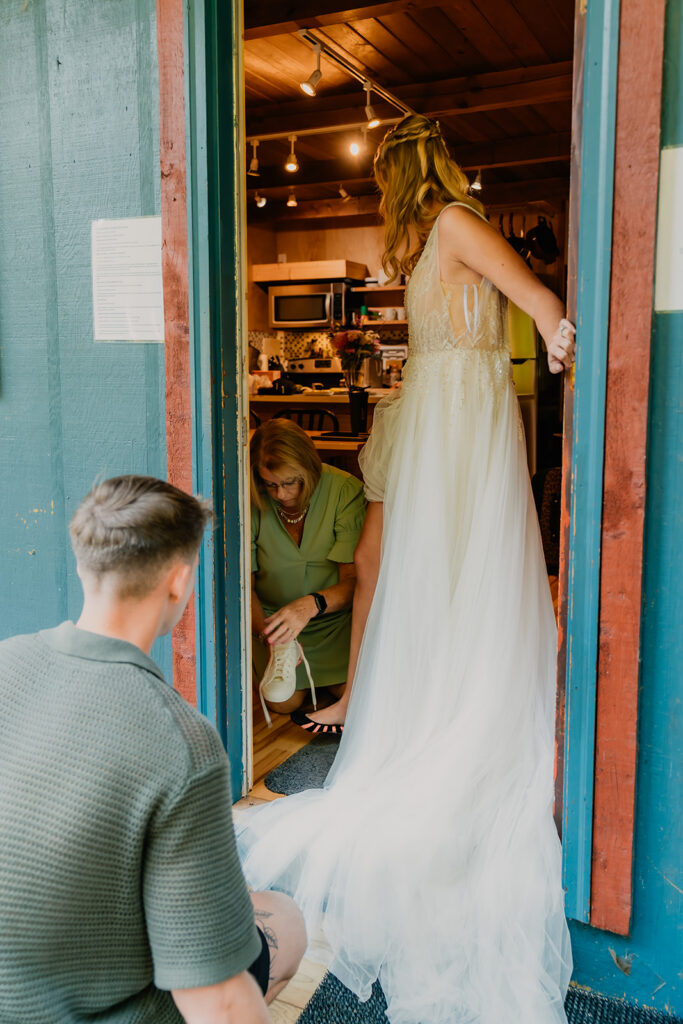 a bride stands in a door frame in her wedding dress. two family members help her get into her shoes to get ready for her mt baker micro wedding 