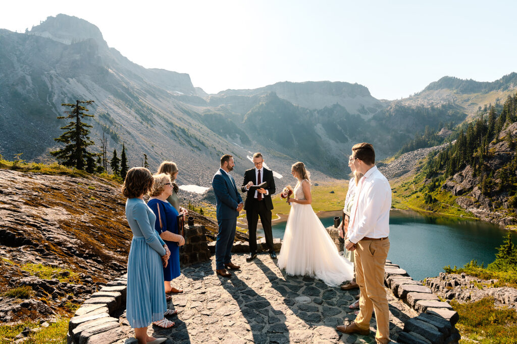 a wedding party stands beside a bride and groom above a sapphire blue alpine lake. the sun shines behind them, we can see that there is a slight breeze in the air during their mount baker micro wedding 