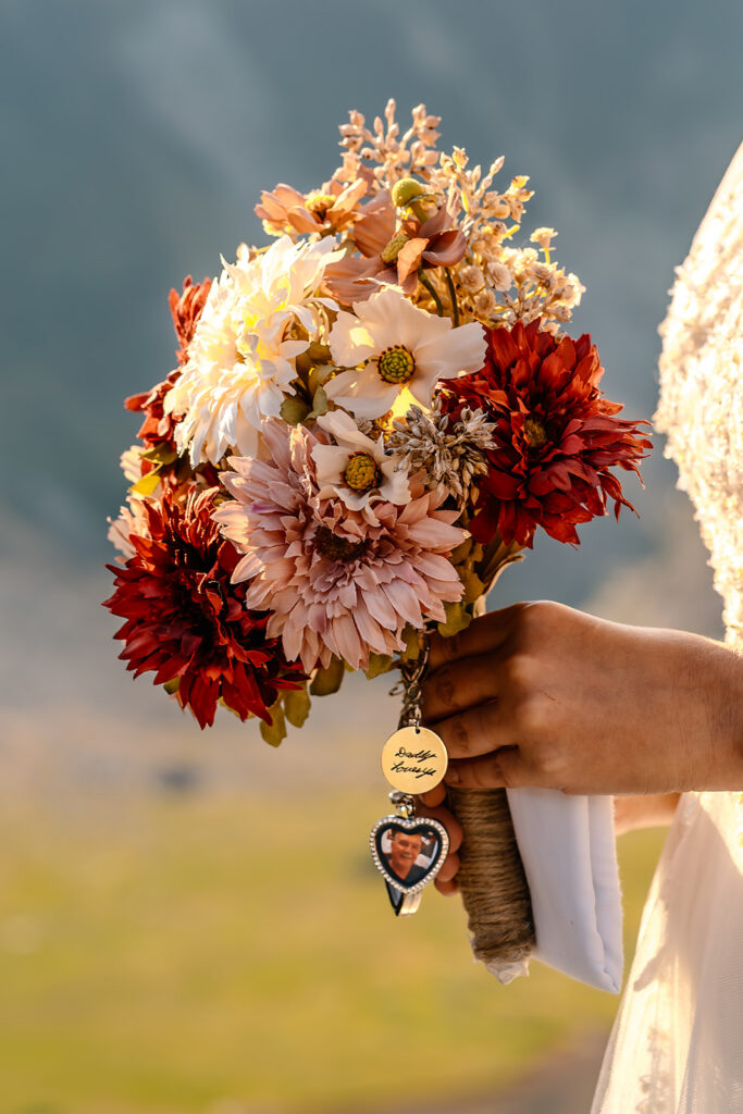 a close up shot of a brides bouquet of blush and bright red flowers. dangling from her bouquet is an inspirited charm and a cherished photo, paying homage to her late father. 