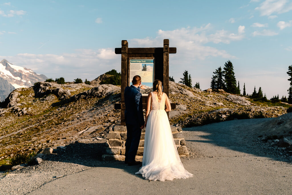 a bride and groom take in a trail sign before embarking on an adventure for their mt baker micro wedding. The sun is shining brightly and the sky is a bright robins blue 